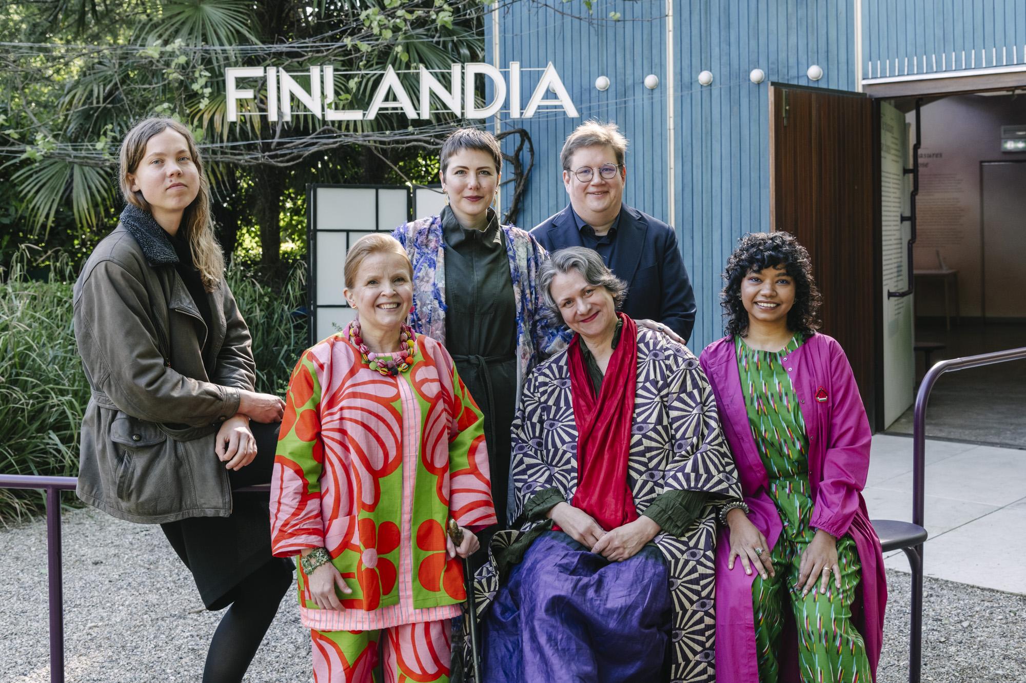 Kuusi ihmistä hymyilee sinisen puisen paviljongin edessä jonka ulkoseinässä lukee isoin valkoisin kirjaimin 'Finlandia'.