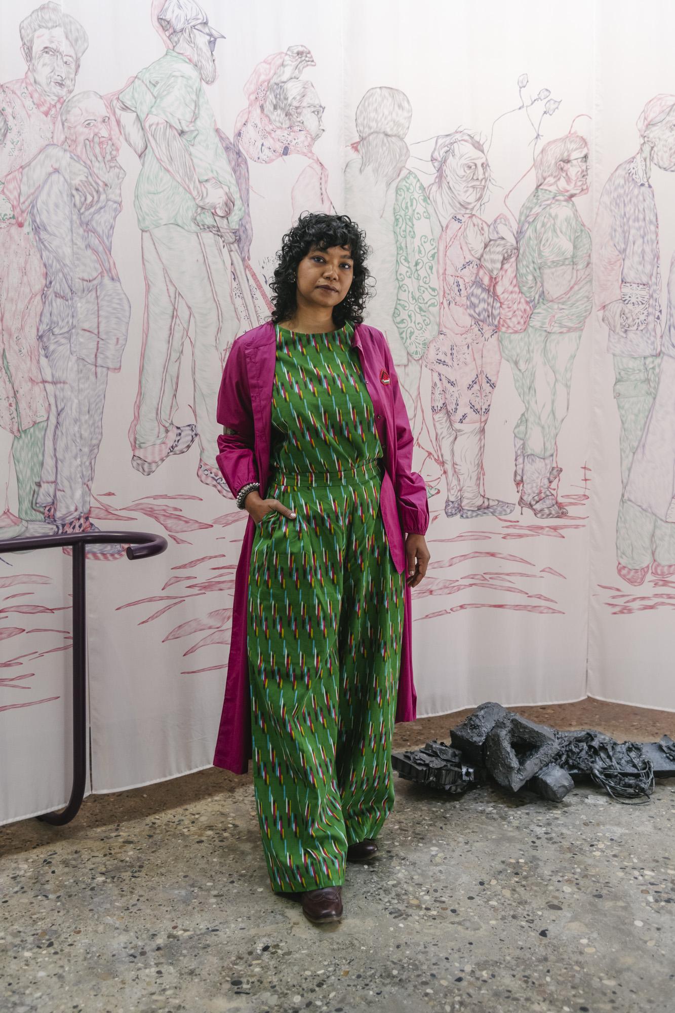 Taiteilija Vidha Saumya pukeutuneena vihreään haalariin ja pinkkiin takkiin seisoo koko taustan peittävän piirroksen edessä, jossa ihmishahmot on kuvattu jonossa.