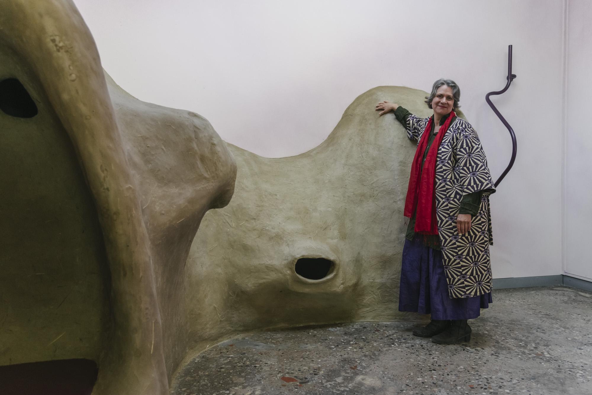 A large beige clay sculpture in an exhibition space, with artist Pia Lindman standing in front of it, wearing a patterned coat and red scarf.