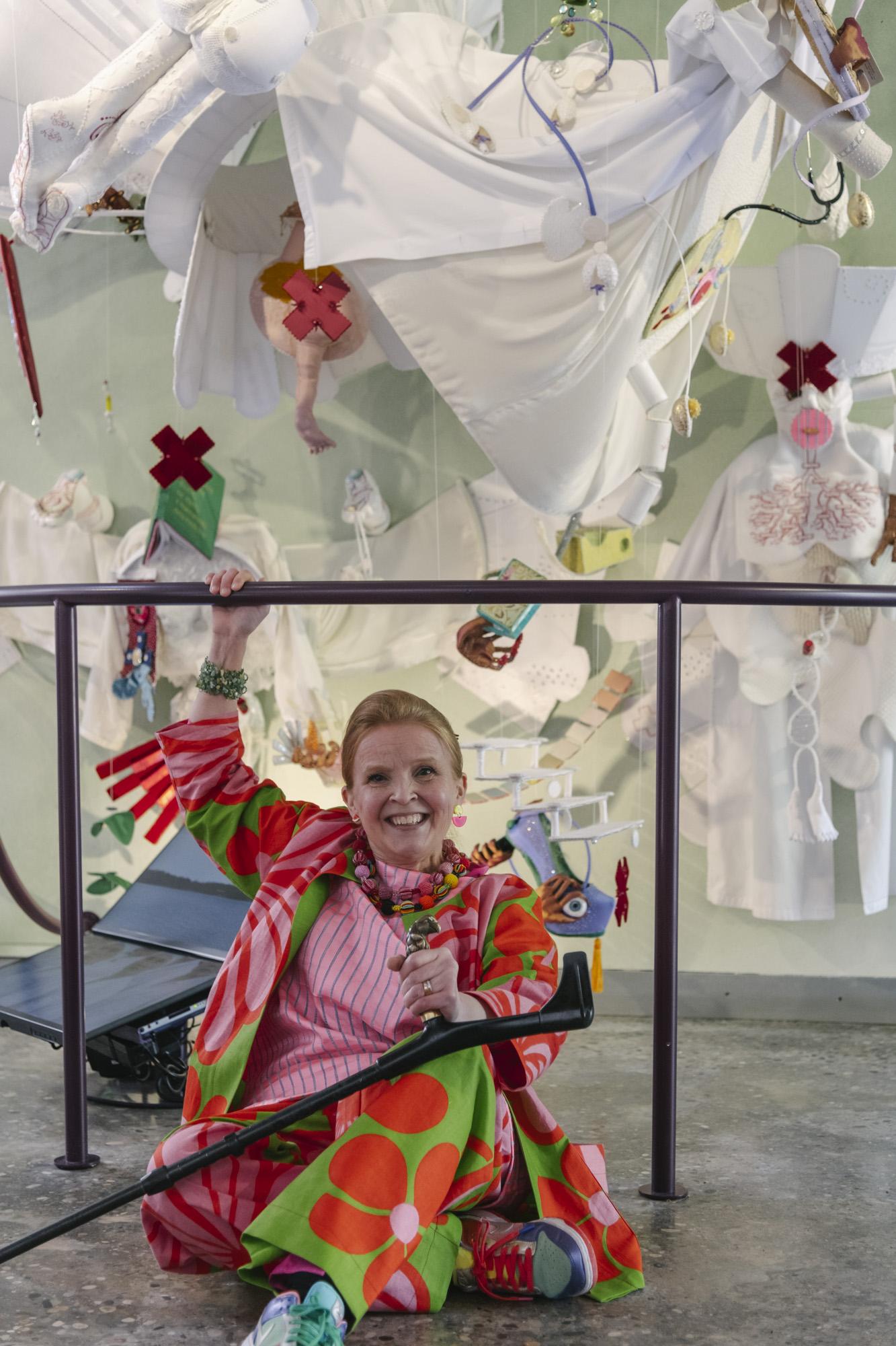Artist Jenni-Juulia Wallinheimo-Heimonen sitting on the floor in front of a large white textile installation with a lot of colorful details. Jenni-Juulia is wearing a red and green suit with patterns and holding a cane.