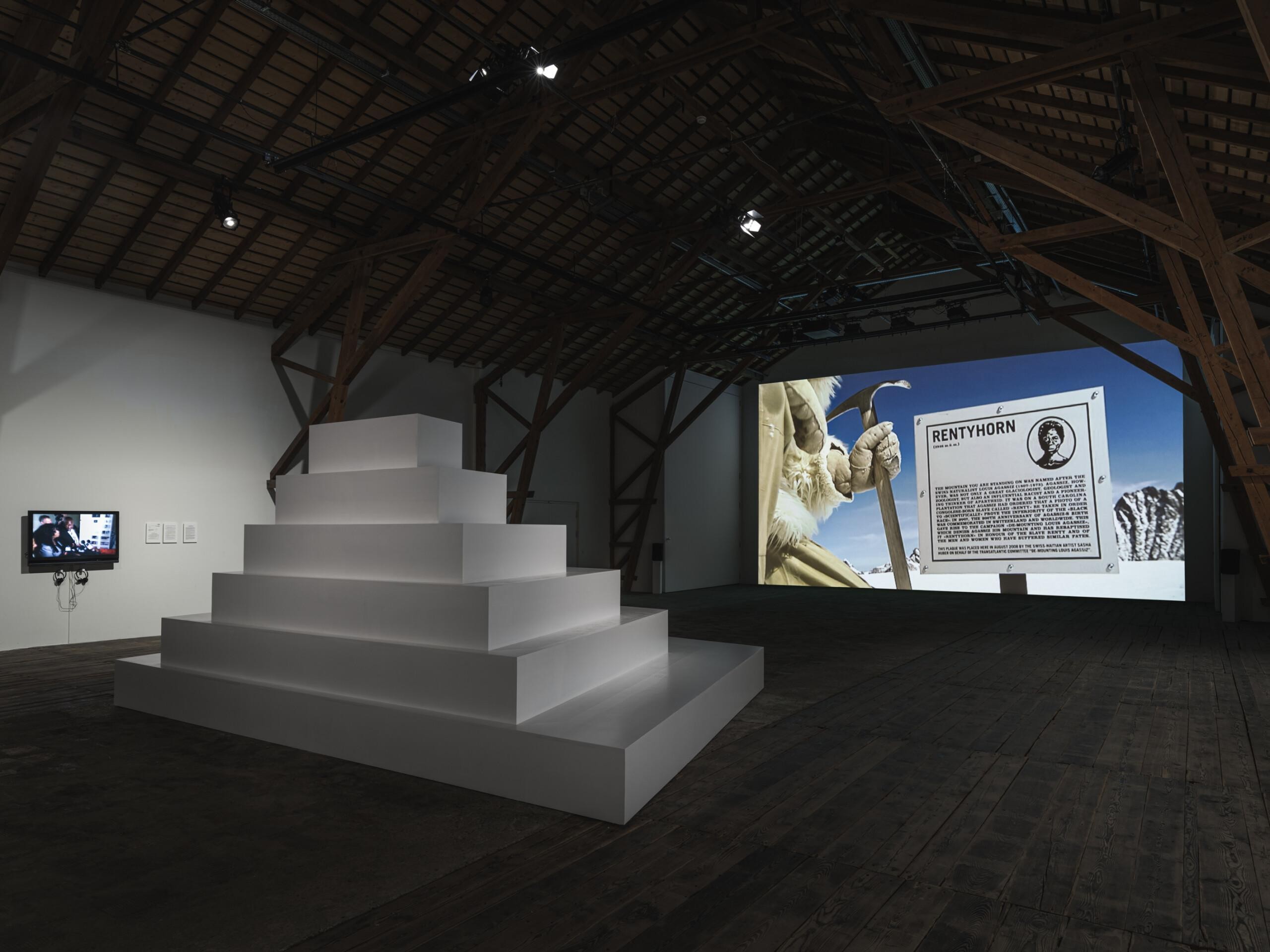 An exhibition space with dark wooden floor and ceiling and white walls. There is a white pyramid-like staircase in the middle of the space, and a big screen in the back wall showcasing art works by artist Sasha Huber.