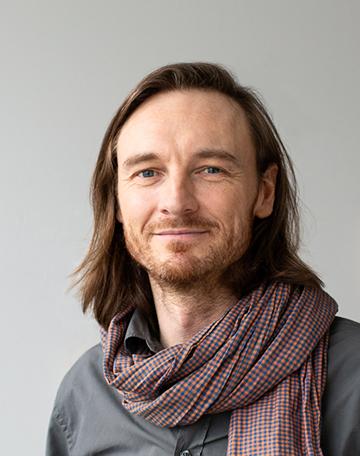 Muotokuva taiteilija James Prevetista, jolla on pitkät ruskeat hiukset, harmaa paita ja ruskea kaulahuivi.