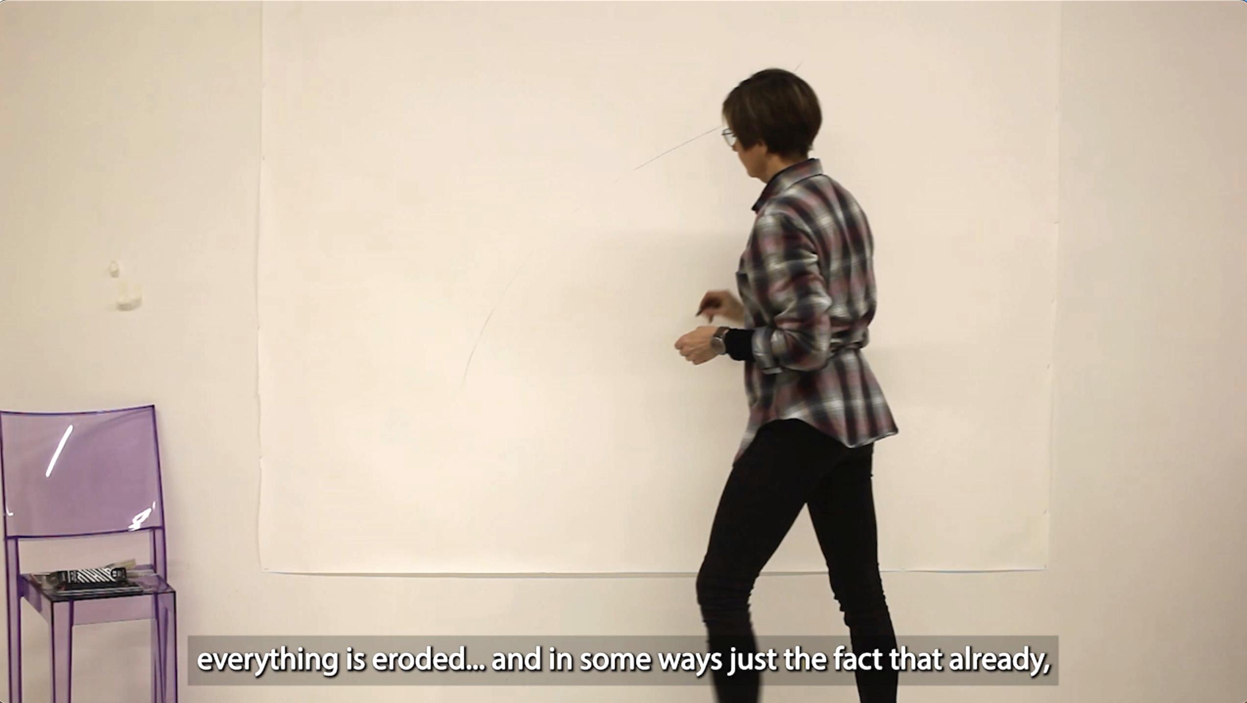 A still from a video where a person is standing next to a white wall with a large paper in it. The person is wearing a purple flannel shirt and black jeans.