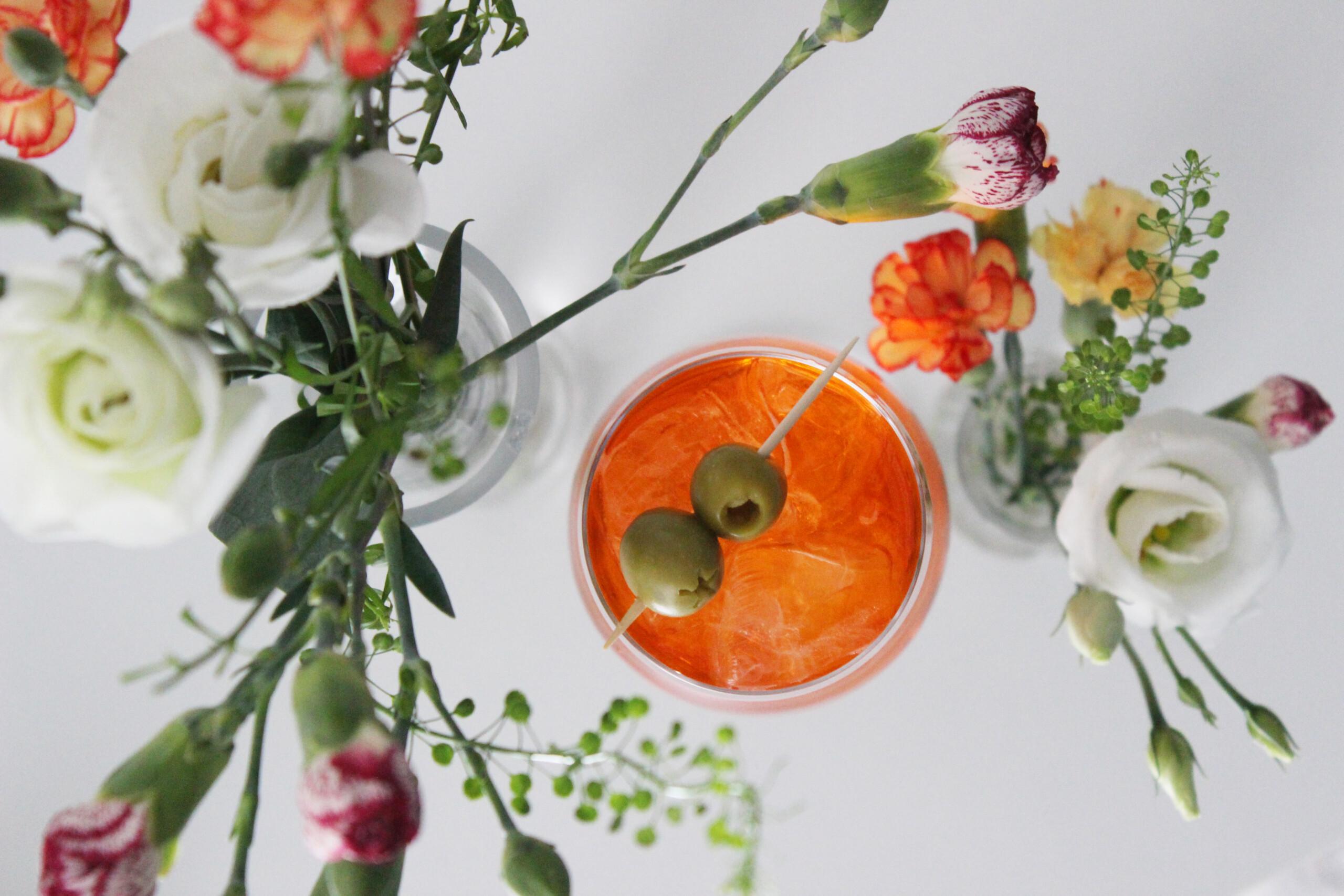 A glass filled with orange drink and two olives on top, viewed from above. There are two glass vases filled with flowers on either side of the glass.