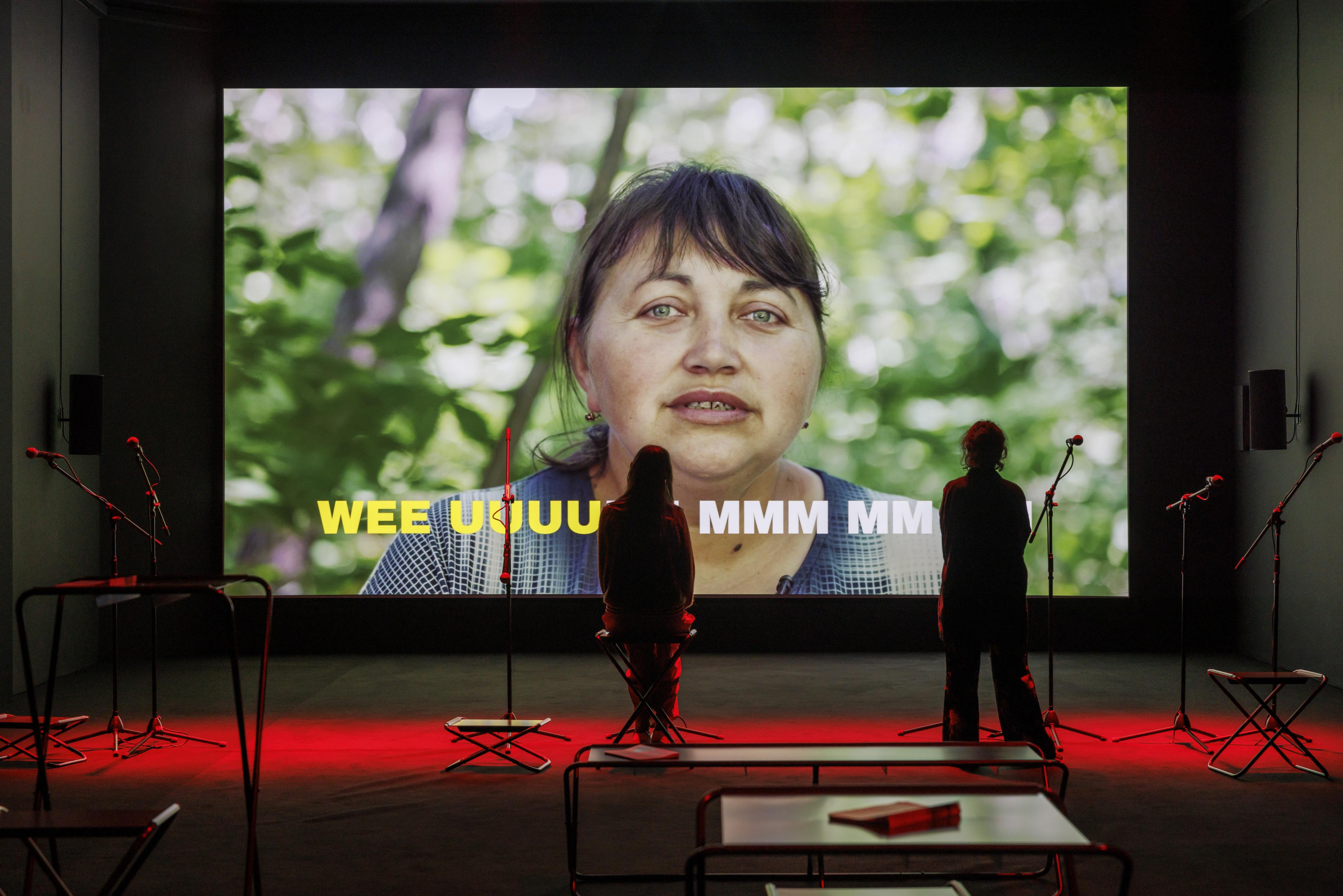 An exhibition space with microphones and a video screen, showing a woman making sounds with her mouth.