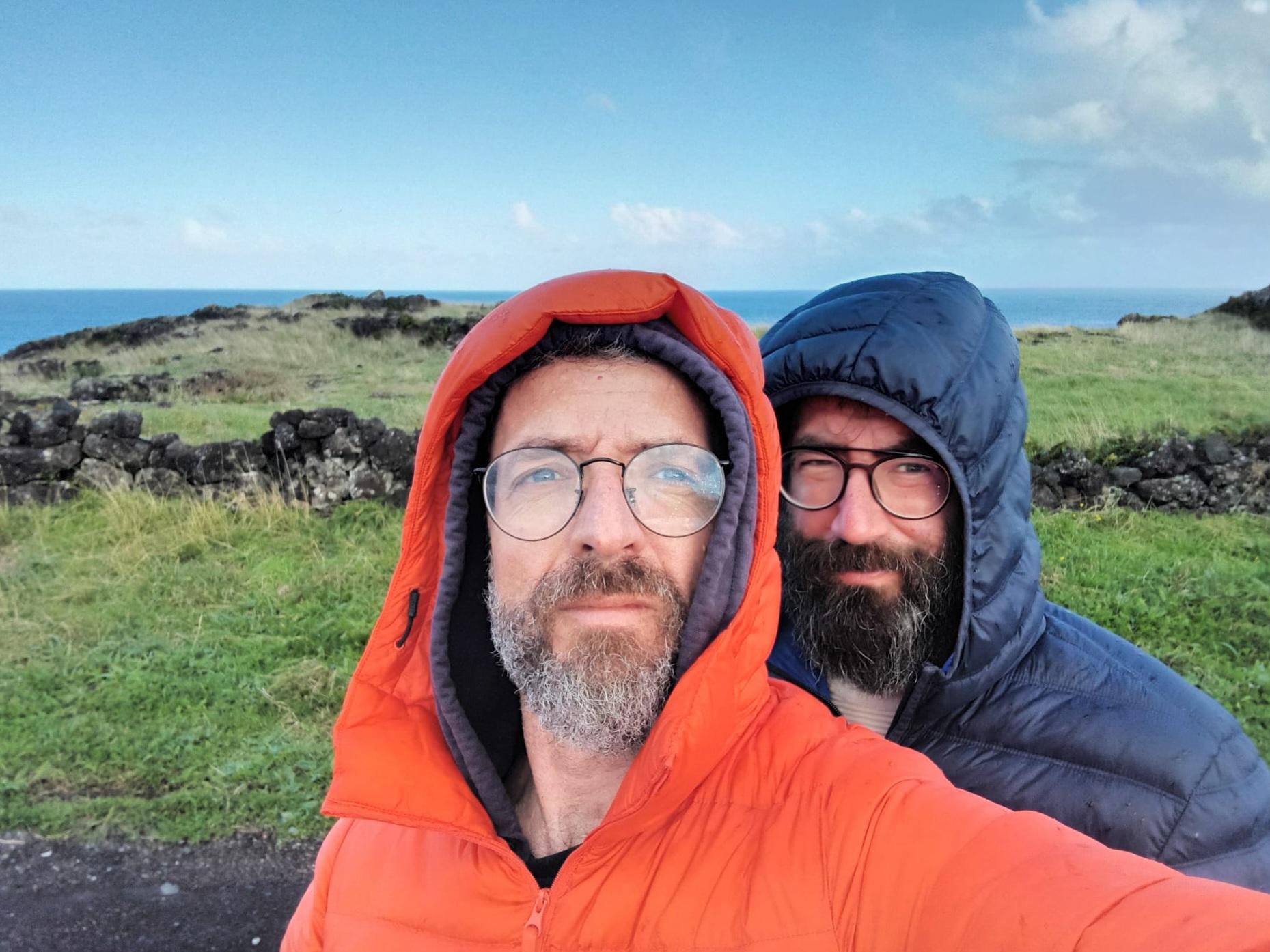 Portrait of two light skinned persons in a open green field with water in the background and a blue sky. Both persons have glasses and facial hair. The person in the front is wearing a red hooded jacket and the person in the back a blue hooded jacket.