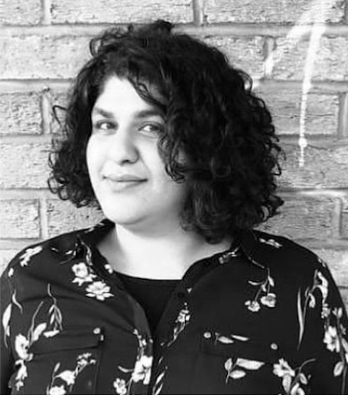 A light skinned person with half long wavy hair wearing a dark flowery shirt in front of a brickwall.