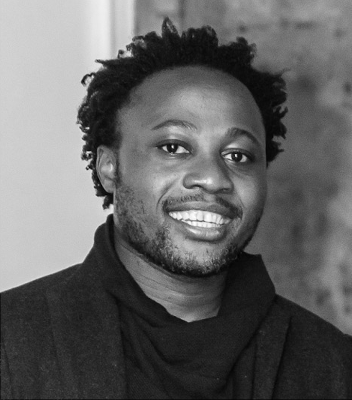 A portrait of a person with brown skin and dark short hair and some light facial hair, wearing a dark shirt and jacket.