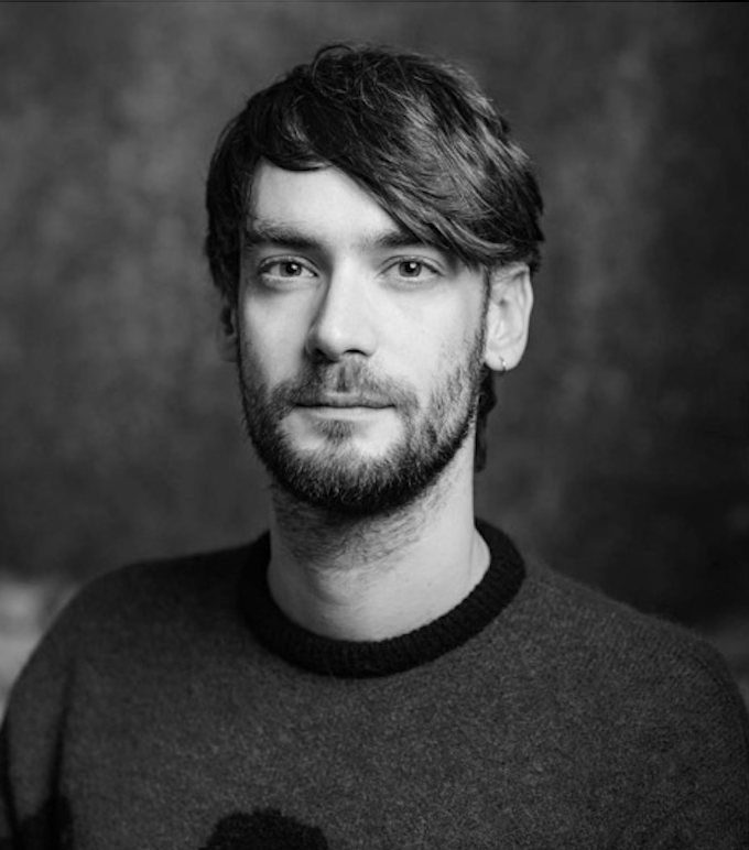 Portrait of a light skinned person with dark short hair and light moustache and beard, wearing a dark shirt in front of a dark background.