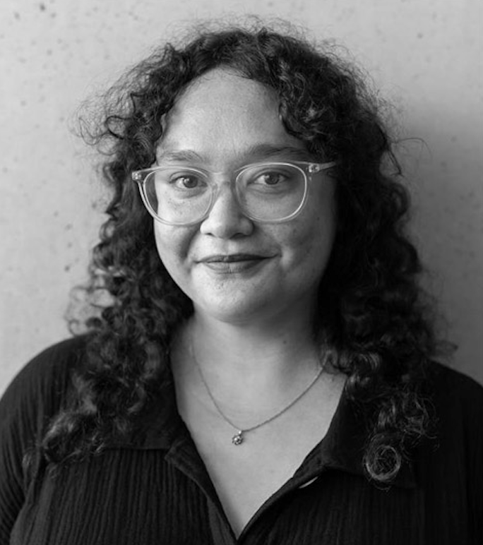 Portrait of a light skinned person with shoulder long dark curly hair, wearing light framed glasses a necklace and a dark shirt with a v-collar.