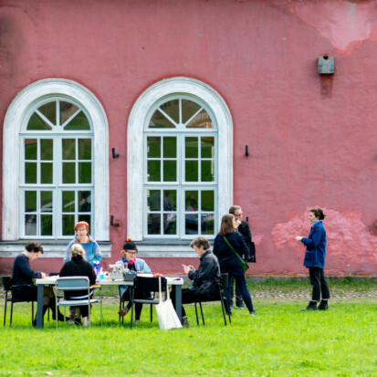 HIAP Suomenlinna premises in the summer. In front of a pink building, people are sitting around a table placed on bright green grass.