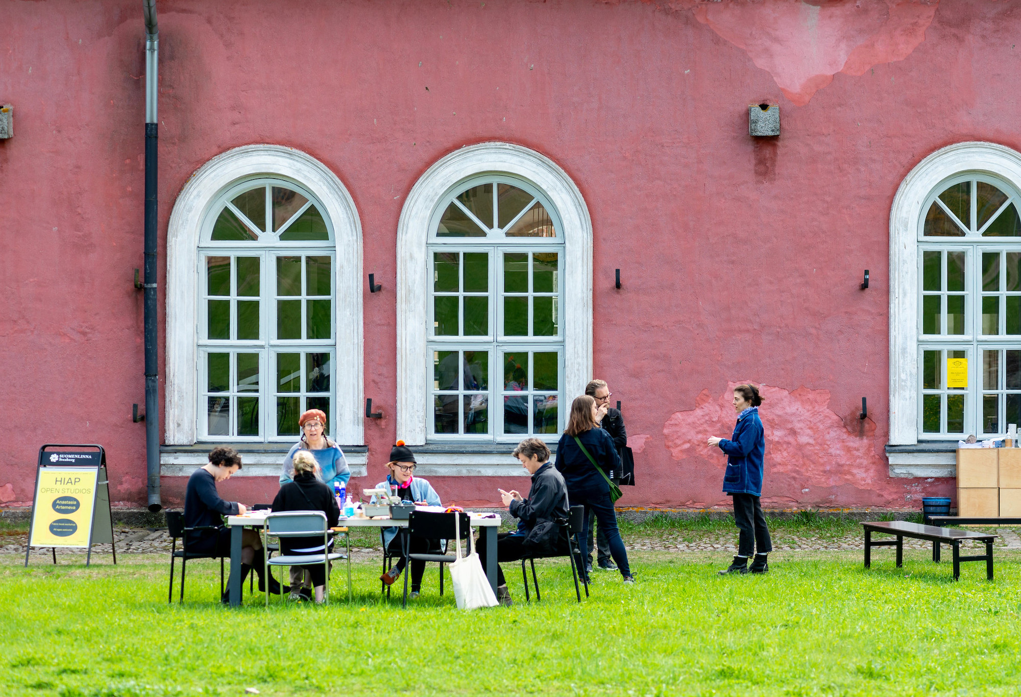 HIAP Suomenlinna premises in the summer. In front of a pink building, people are sitting around a table placed on bright green grass.