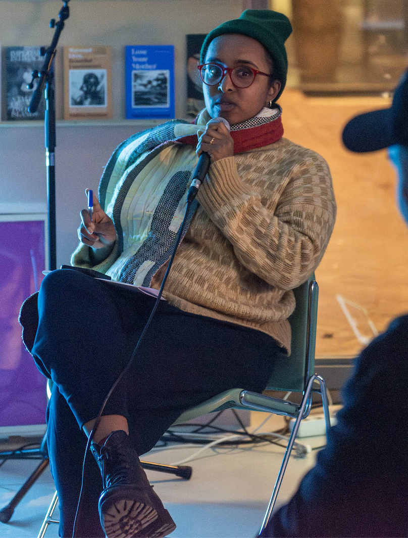 Amal sits in the space and holds the microphone during an event at Metro54, in conversation with guest speakers. Audience members are present in the space.