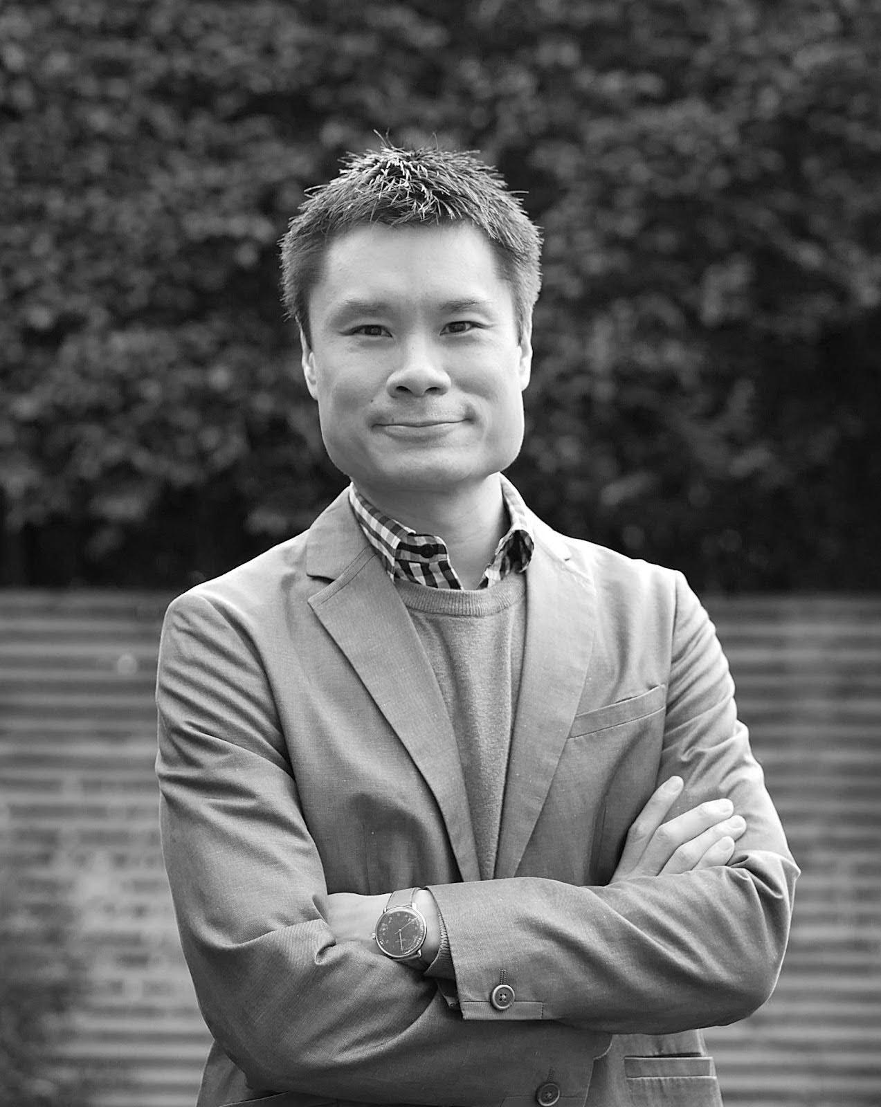 Cliff Lauson Black and white portrait of Cliff Lauson in a blazer with arms crossed, standing outdoors and smiling against a blurred greenery backdrop.